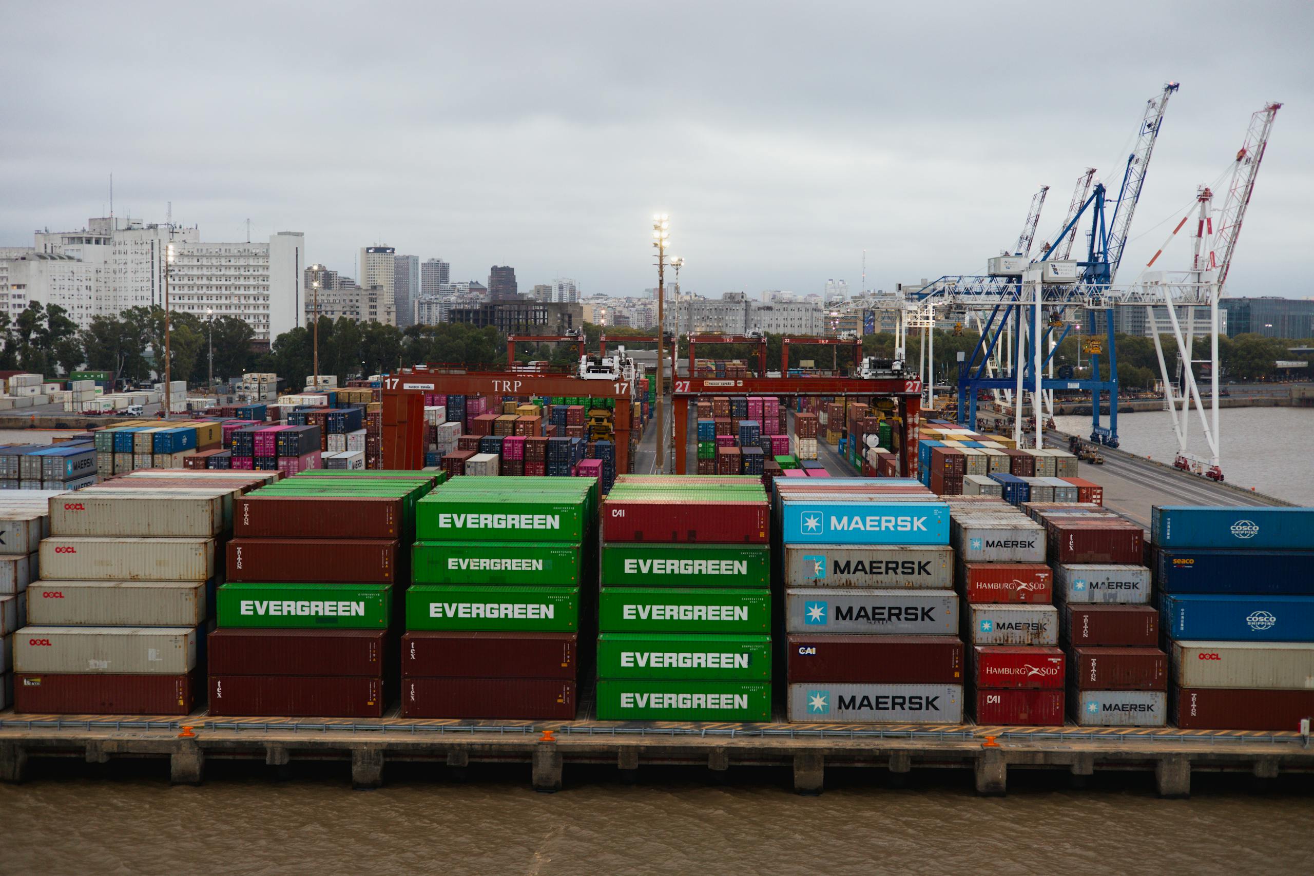 Aerial view of colorful shipping containers in a bustling port with cranes and city skyline.