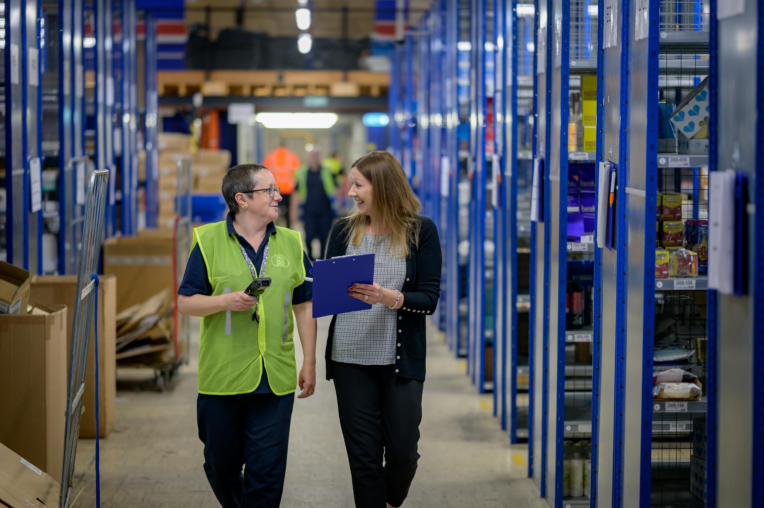 Two workers in reflective vests collaborate in a busy warehouse aisle.